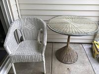 White wicker armchair next to round wicker table with glass top on a concrete porch.