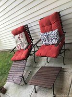 Two outdoor lounge chairs with red cushions, wooden slat backs and seats, each paired with a detachable wooden slat leg rest. One chair has a floral pillow, the other a paisley pillow.