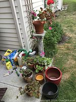 Live outdoor plants in pots, concrete plant stand, gardening pots, watering can, and gardening supplies all arranged on a concrete patio area near a garden bed.