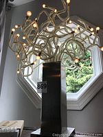 Contemporary table lamp shown from side angle in front of window, displaying its multiple transparent acrylic branches with small round light bulbs at tips and rectangular metal base.