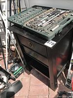Socket and bit set in green plastic case on top of black wooden storage table with two drawers and open shelf.