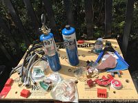 Photo showing two blue propane torch cylinders with valves and hoses on a wooden work surface along with various copper plumbing connectors, pipe cutter, and other assorted tools and materials.
