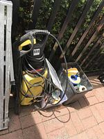 Yellow Karcher power washer with hoses and power cable leaning against a fence next to a black bin containing painting supplies.