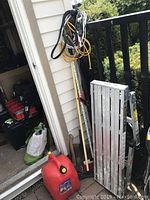Photo shows adjustable folding ladder, coiled extension cords, jumper cables hanging on railing, red plastic gas can on ground, and additional hand tools leaning against wall.