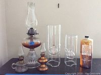 Lot showing all six oil lamps and the lamp oil bottle on a table against a plain wall for size and style comparison.