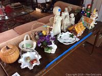Wide view of table with ceramic holiday decor items including Christmas angel candle holder, ceramic houses, pumpkin, Easter basket with eggs and flowers, hand painted eggs, and figurines.