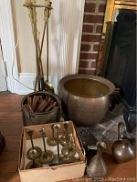 Photo showing brass candlestick holders in a cardboard box, hammered brass pot, metal pot with wood fat sticks, and metal fireplace accessories nearby.