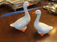 Photo of two white ceramic geese figurines with orange beaks and feet, placed on a wooden table.