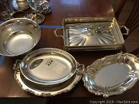Five silverplate serving items displayed on wooden surface: Paul Revere reproduction bowl with lid, covered holder tray without dish, and three serving platters with decorative rims and handles.
