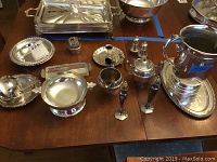 Photo showing overall assortment of silver and silver-plated tableware items on wooden table, including pitchers, bowls, trays, salt and pepper shakers, and candle holder with snuffer.