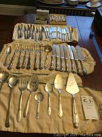 Full view of laid out silver plate flatware and serving pieces on cloth with boxed baby utensils and miniature spoons in the background.
