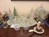 Overview of various crystal and glass pieces including trees, covered bowl, hobnail creamer and sugar bowl, candle holder and mirror tray on dark wood surface.