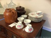 Lot shown on wooden dresser with 2 covered stoneware jars, 6 tea cups, and 8 luncheon plates with floral pink rose decoration.