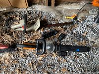 Top view of both hedge trimmers lying on gravel, Worx trimmer bottom, Sears trimmer top, including charger and battery packs near Worx trimmer.
