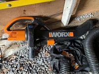 WORX electric leaf blower with black and orange color scheme placed on gravel near a wooden wall.