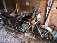 Left front side view of 1974 Yamaha RD350A on wood blocks inside shed showing overall condition, paint and surface rust