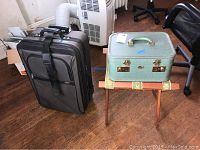 Grey soft-sided suitcase and vintage light green cosmetic case sitting on wooden folding luggage rack on a hardwood floor.