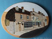 Front view of the hand painted oval wood plaque depicting store fronts on a busy street, showing details of buildings, windows, doors, a flag, and street signs.