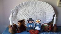 Large white ceramic turkey platter with detailed feather texture, shown with Thanksgiving holiday decorations including Pilgrim figures, turkey candle holders, haystack, pumpkin, and small turkey figurine.