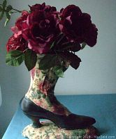 Side view of Victorian style boot vase showing floral fabric upper and black shoe heel base with red flowers inside