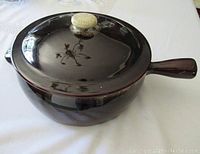 Large brown glazed ceramic bowl with handle and matching lid on, photographed on a white table cloth.