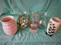 Five large vases placed in a row on a green cloth background, showing varied styles, colors, and decorations including floral motifs and glass patterns, with one signed and one identified as Hull.