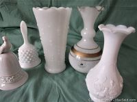 Five milk glass items including two bells and three vases with varying designs and detailing, white milk glass with one vase featuring gold bands.