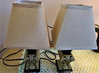 Two table lamps each with beige fabric trapezoid lampshades and wood-like square bases with decorative pattern on front. Electrical cords visible behind.