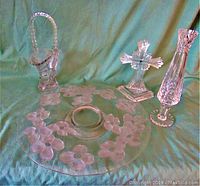 Photo showing frosted glass cake plate with floral design, crystal cross figurine, tall cut crystal vase and bride's basket arranged on green fabric.