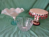 Three items on green fabric background: ruffled milk glass bowl on green pedestal, clear crystal bowl, and decorative cake dish with cherry pattern and red bead accents.