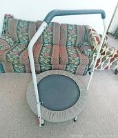 Mini exercise trampoline rebounder with white frame, beige padding, and black jumping mat shown in front of a patterned couch
