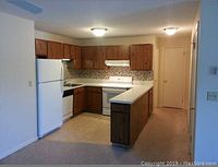 Photo shows a U-shaped kitchen area featuring medium brown wood-grain laminate cabinets above and below the countertop, white stove and refrigerator, mosaic tile backsplash, and beige vinyl flooring.