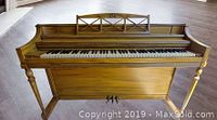 Front view of wooden piano showing full keyboard, carved music stand, legs, and pedals on wooden floor.
