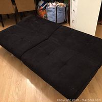 Black futon mattress laid out flat on a wood floor. Shows wear, fabric texture, and condition.