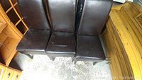 Photo showing three brown leather dining chairs arranged side-by-side. Leather surfaces showing wear and scuff marks, but padding looking intact. Wooden legs visible with dark finish.