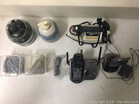 Photo of a household lot containing an electric iron, two cordless telephones with bases and handsets, two containers of large silver-tone paper clips, a container of decorative polished gray stones, and a ceramic soap dispenser with pump.