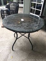 Rattan patio table with round glass top on black metal stand seen from side angle. Worn rattan weave under clear glass surface.