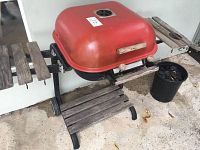 Photo showing the red charcoal grill with wooden side and bottom shelves, two wheels, and a vented lid.