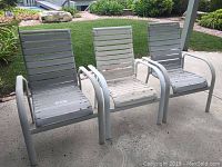 Three white outdoor patio chairs with metal slatted seats and backs and curved armrests placed on concrete patio.