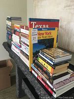 Books stacked along a surface including atlases for Texas and New York and several other books mostly paperback and some hardcover