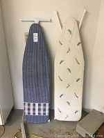 Two full-sized ironing boards side by side against a wall, one with a blue pinstripe cover and one with a feather print cover.