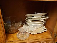 Photo showing stacked white ceramic bowls and platters alongside clear glass bowls and a two-tier glass serving stand inside a wooden cabinet.