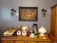 View of decorative ceramic and glassware items on wooden surface underneath the metal artwork, including vase, black floral vase, serving bowl with lid, glass jars, gold-trimmed glasses, cup and saucer, tray, and glasses