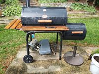 Full view of Char-Griller black charcoal grill and smoker with wooden handles and side shelves, standing on wheels on patio, with some stored items underneath