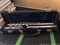 Open black hard flute case labeled Armstrong containing a disassembled silver flute resting in blue velvet interior.