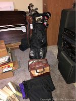 View of two golf bags side by side with golf clubs inside, and the bowling ball bag on the floor near some books.