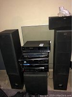 Front view of stacked electronics including Panasonic VHS players, Sony DVD player and Sony audio receiver gear between two tall black Sony tower speakers and a subwoofer on floor
