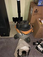 Front view of the Ridgid Wet Vac model WD06250 showing the unit with attached power cord, black hose sections mounted vertically on wall, and filter sleeve on floor, surrounded by various boxes.
