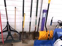 Photo showing multiple large plastic yard rakes, four metal shovels with wooden handles of different shapes, a yellow plastic snow shovel blade, and a blue backpack sprayer.