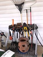 Photo showing garden tool caddy loaded with various rusted gardening tools including rakes, hammers, axes, pruners, and hand tools along with a terracotta chipped garden pot at the base.